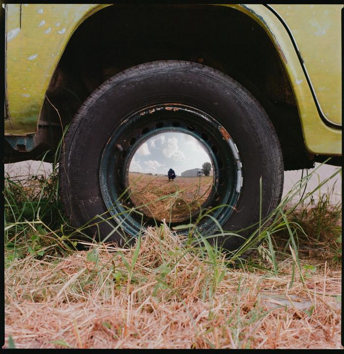 A person is reflected in the hubcap of a vehicle parked in a field.