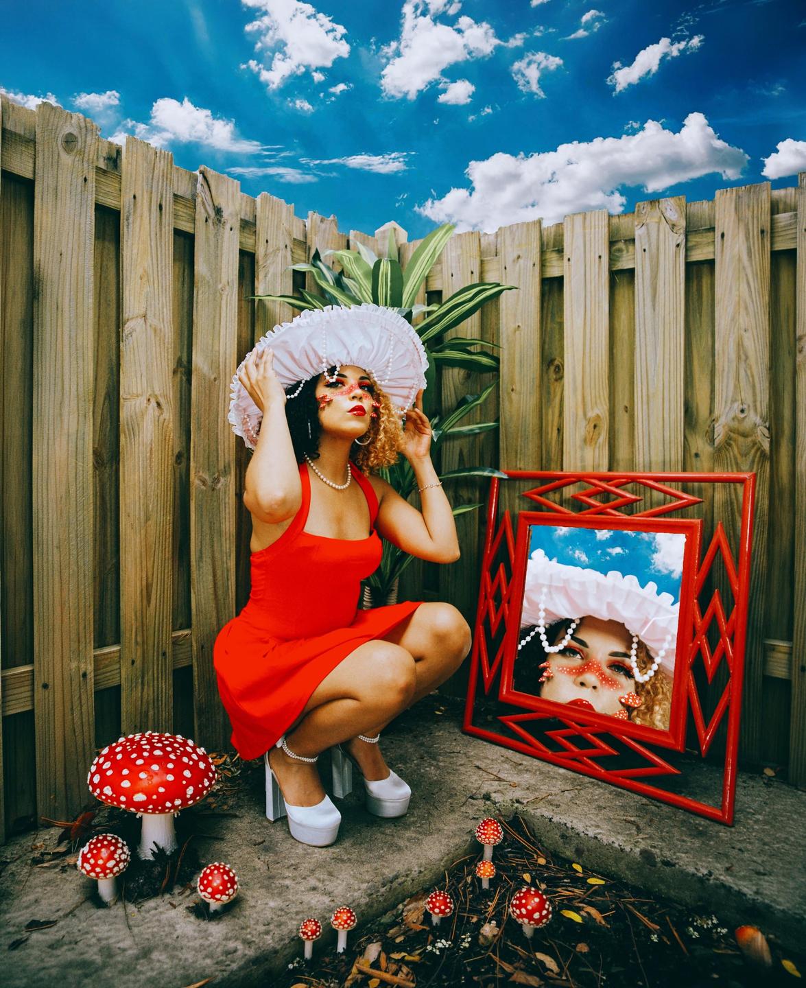 A woman in a red dress and white hat crouches next to a mirror and red and white mushrooms.