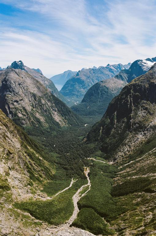 A deep valley with a river running through a forest is surrounded by mountains under a blue sky.
