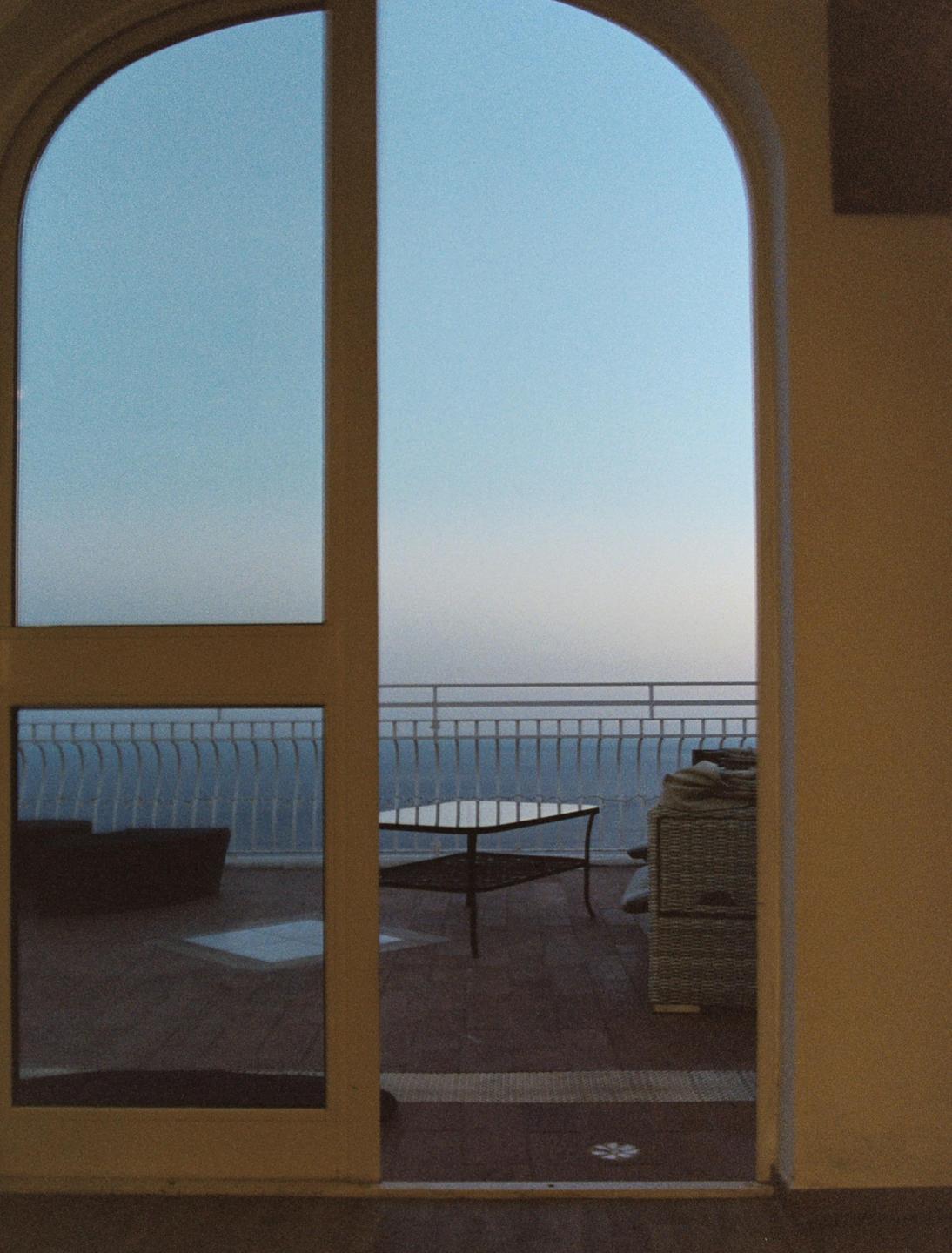 A view of a balcony with a table and chairs overlooking the ocean is seen through an arched doorway.