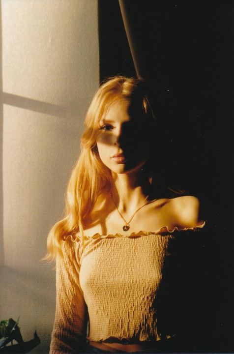 A young woman with long blonde hair wears an off-the-shoulder top and a necklace, with her face partially in shadow.