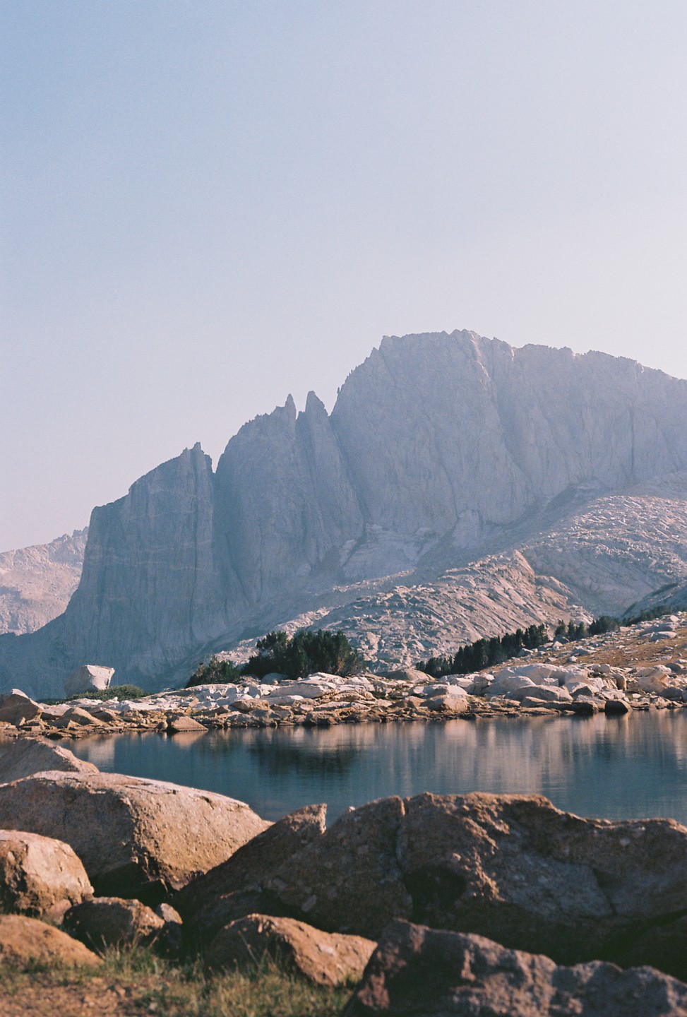 A large mountain range rises behind a calm lake with rocks in the foreground.