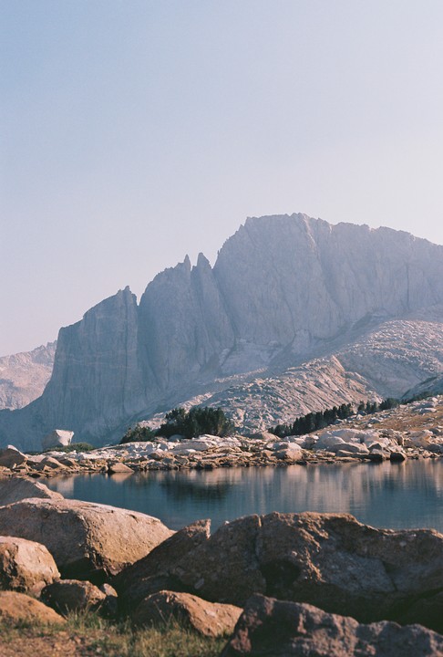 A large mountain range rises behind a calm lake with rocks in the foreground.