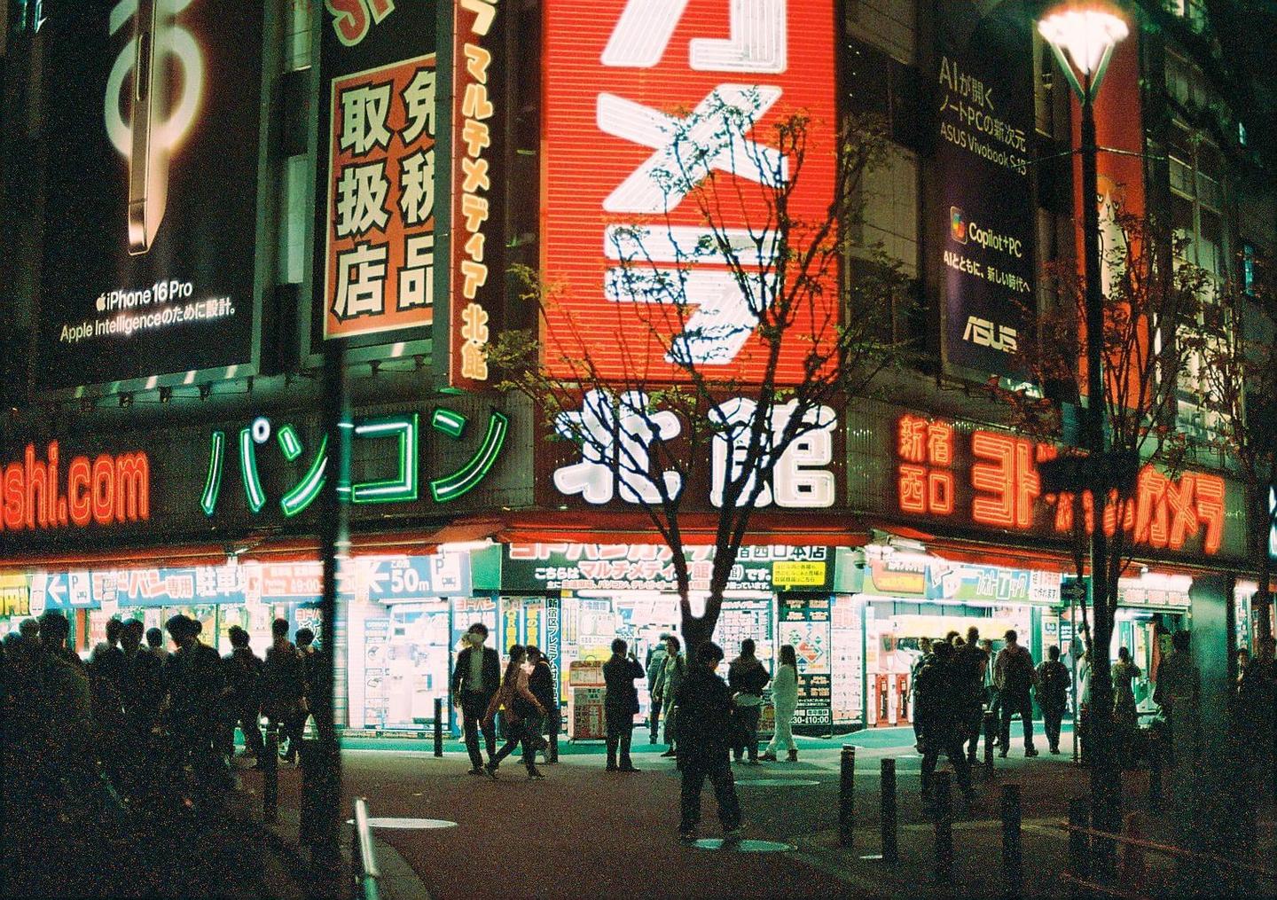 A street in Tokyo is bustling with people walking past buildings adorned with bright neon signs at night.