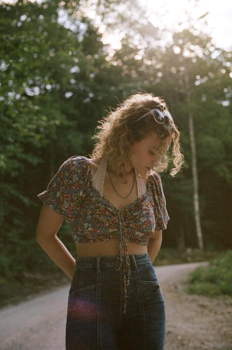 A woman with curly hair and sunglasses on her head stands on a dirt road, looking down.