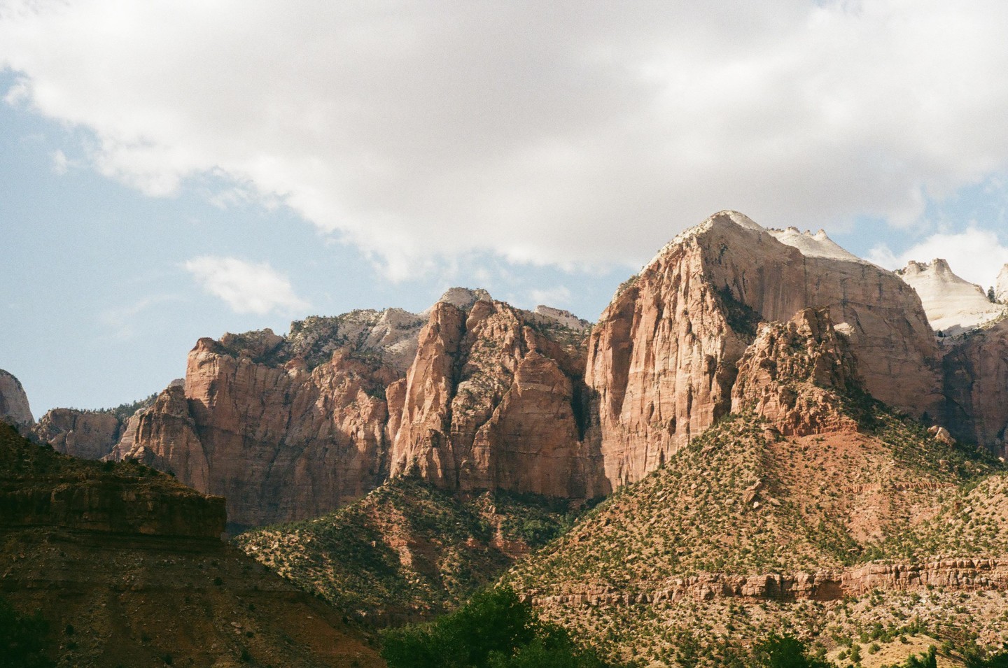 A vast landscape shows towering red rock mountains covered in sparse green vegetation under a partly cloudy sky.