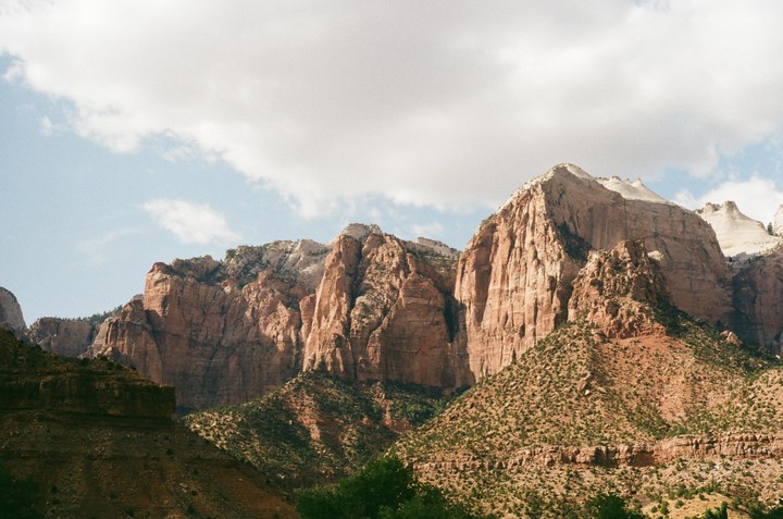 A vast landscape shows towering red rock mountains covered in sparse green vegetation under a partly cloudy sky.