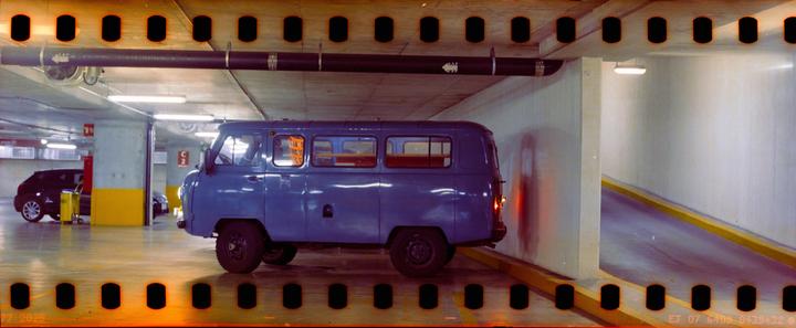 A blue van is parked in an underground parking garage next to a concrete wall and a yellow curb.