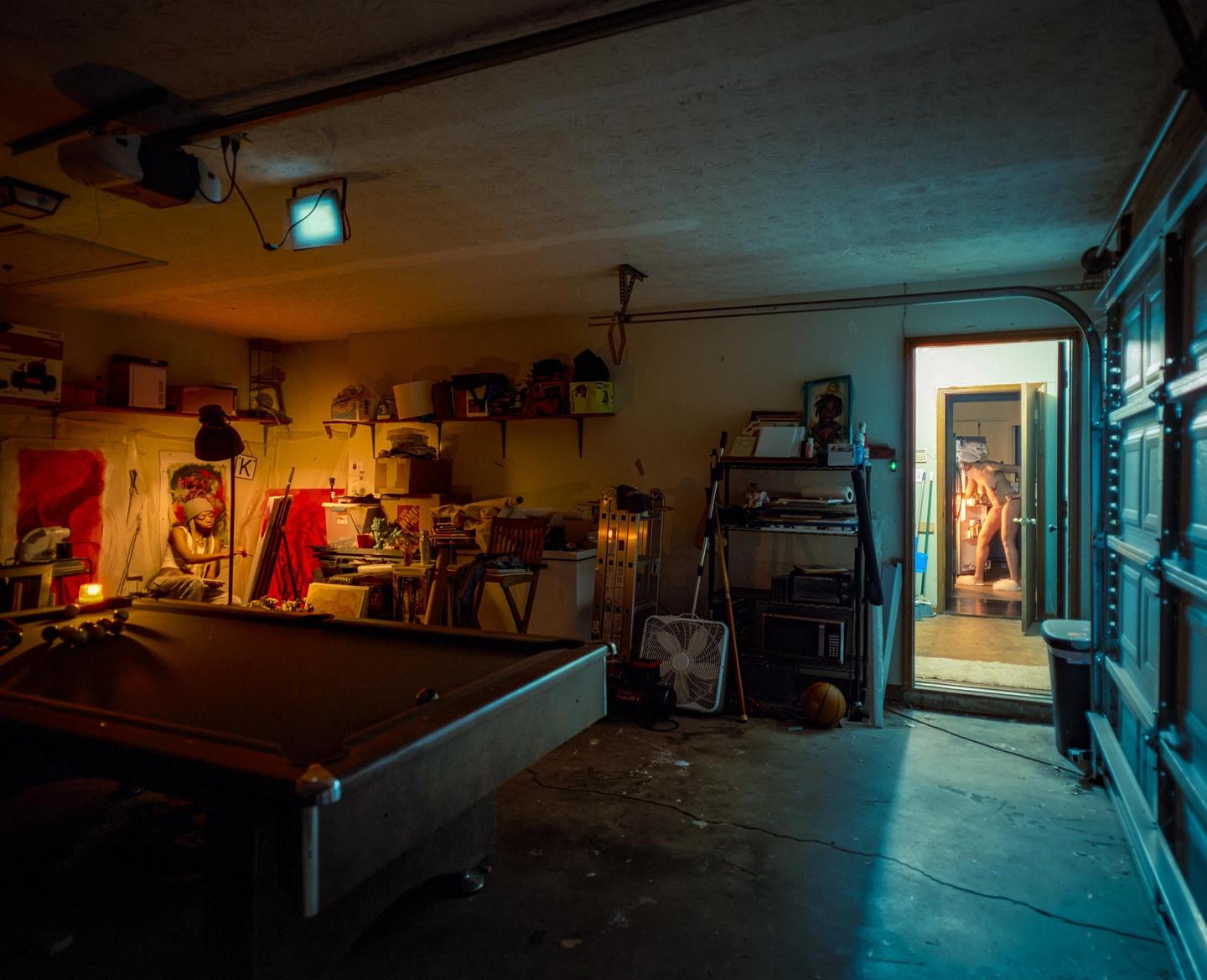A woman sits in a cluttered garage with a pool table while another woman stands in a doorway in the background.