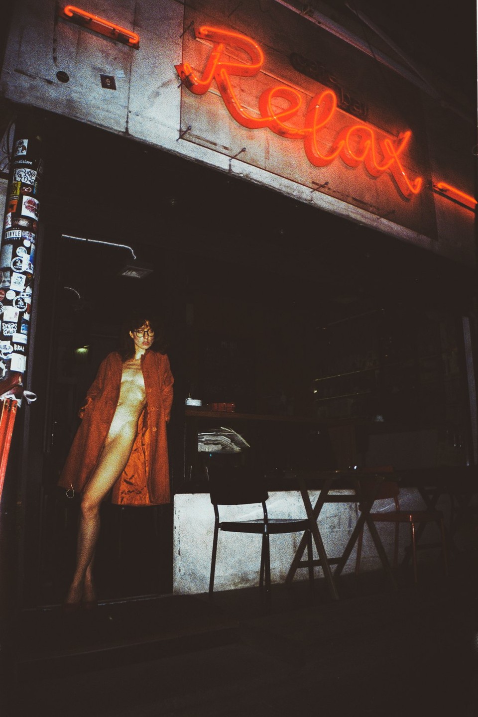 A woman in a red coat stands in a doorway under a neon sign that says "Relax" at night.