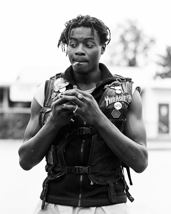 A man with dreadlocks and a vest with patches holds a lighter to a cigarette in his mouth.