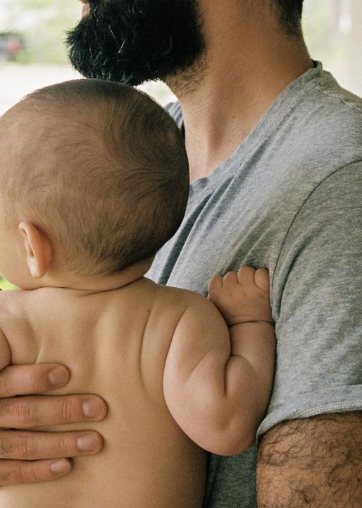 A man holds a baby with the baby's back to the viewer.