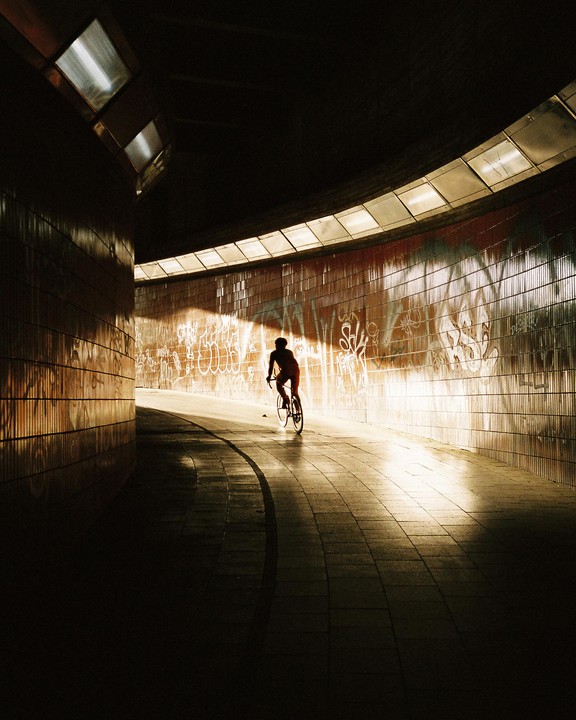 A cyclist rides through a tunnel with graffiti on the walls, illuminated by a bright light source ahead.