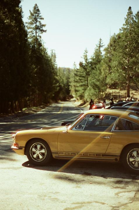 A yellow Porsche is parked on the side of a road lined with tall trees.