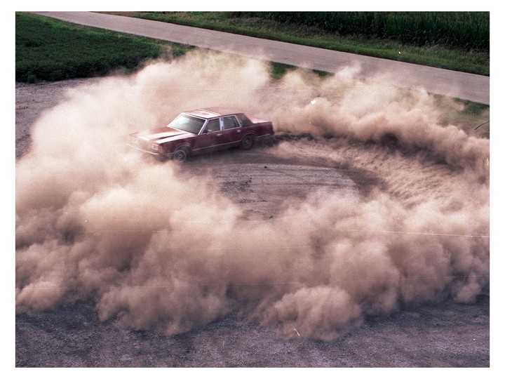 A red car kicks up a large cloud of dust as it turns in a circle on a dirt road.