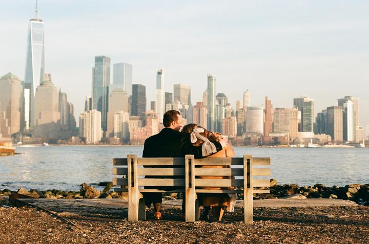 A couple sits on a bench facing the new york city skyline across the water.