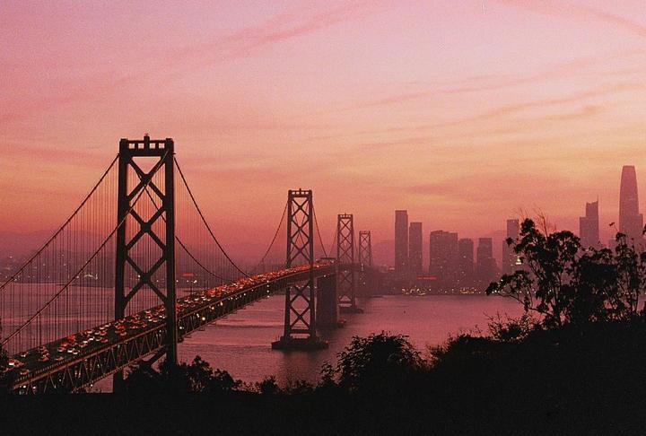 A suspension bridge with heavy traffic crosses a body of water towards a city skyline under a pink and orange sunset.