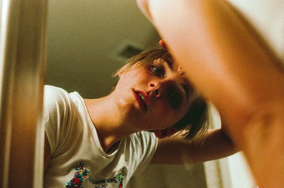 A young woman with short hair looks into a mirror, with her reflection visible.