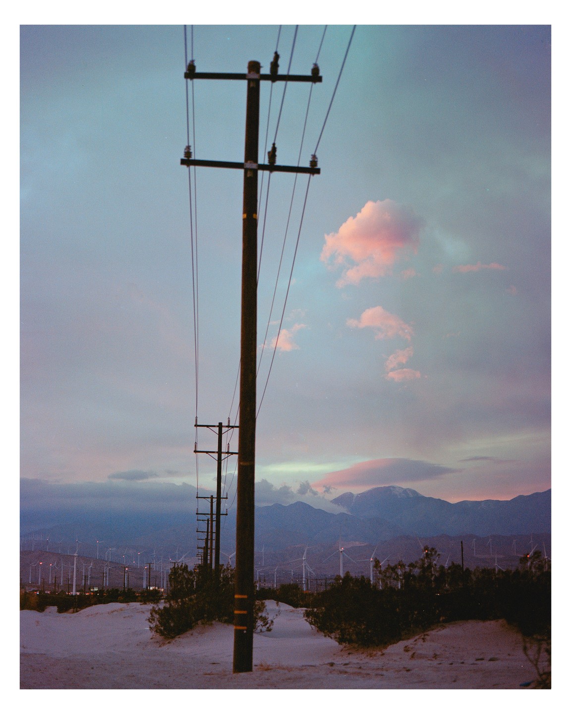 A row of utility poles stands in a desert landscape with mountains and wind turbines in the distance under a colorful sky.