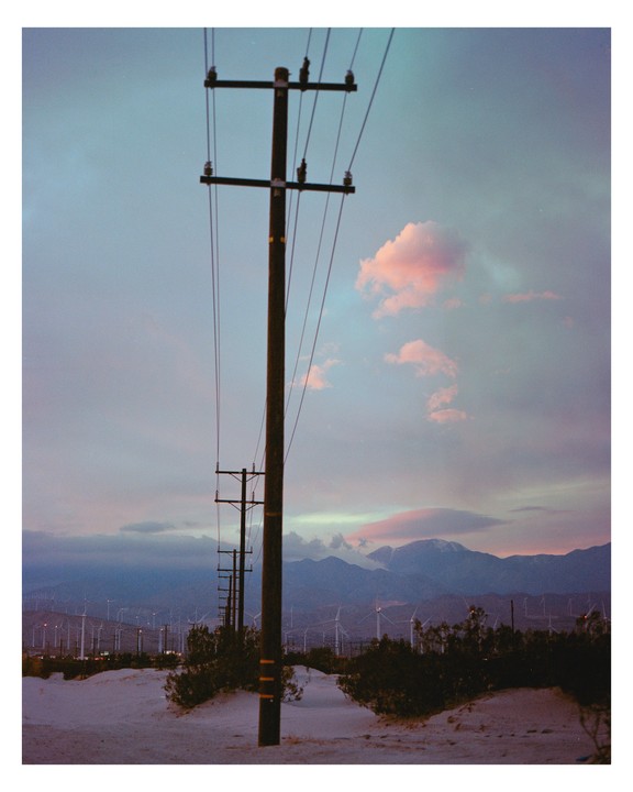A row of utility poles stands in a desert landscape with mountains and wind turbines in the distance under a colorful sky.