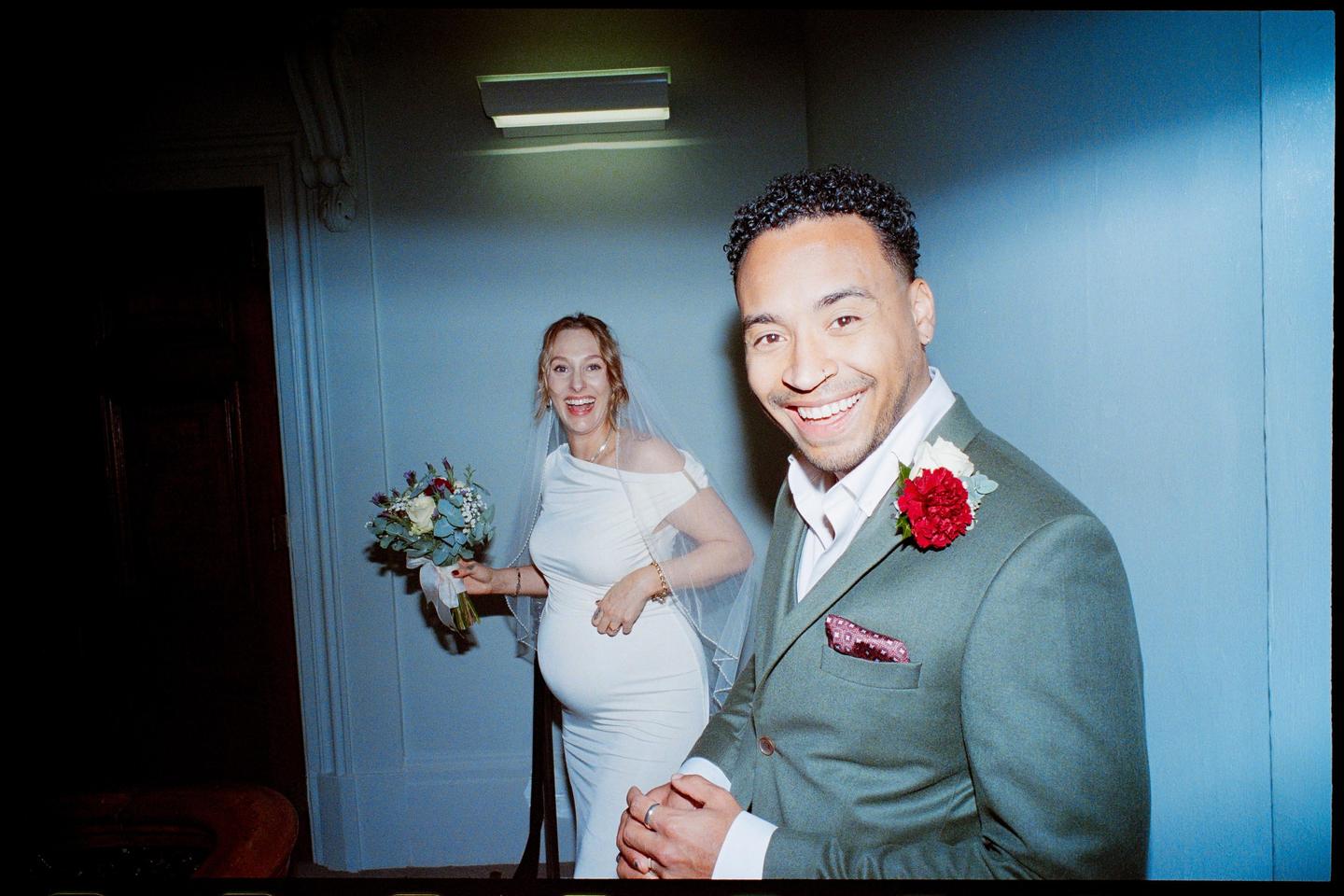 A man in a suit and a pregnant woman in a wedding dress smile at the camera.