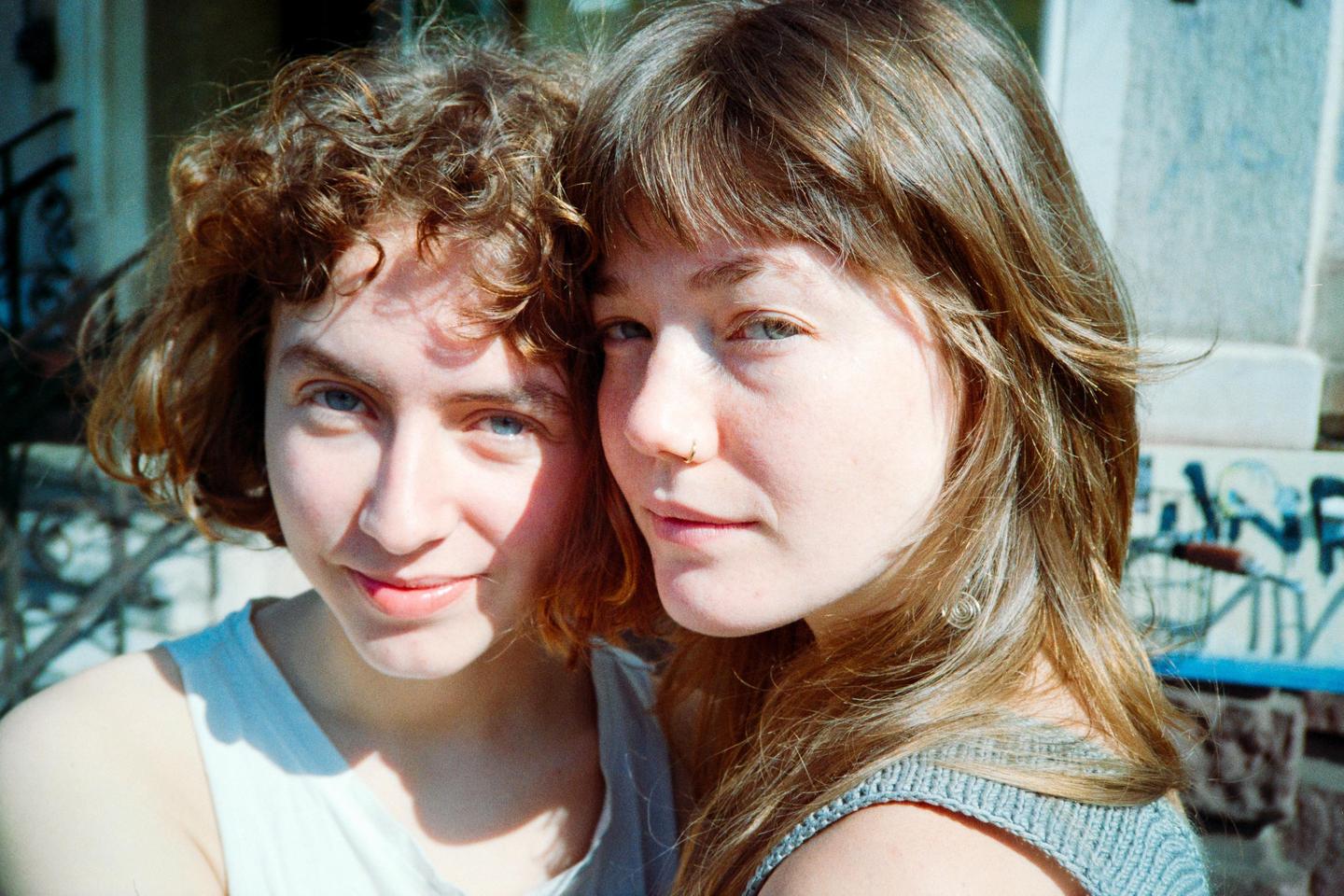 Two women are posing for a close-up portrait outdoors with graffiti in the background.