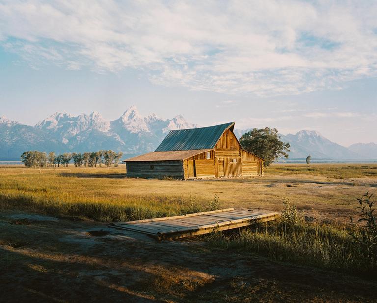 A wooden barn stands in a field with mountains in the background under a cloudy sky.