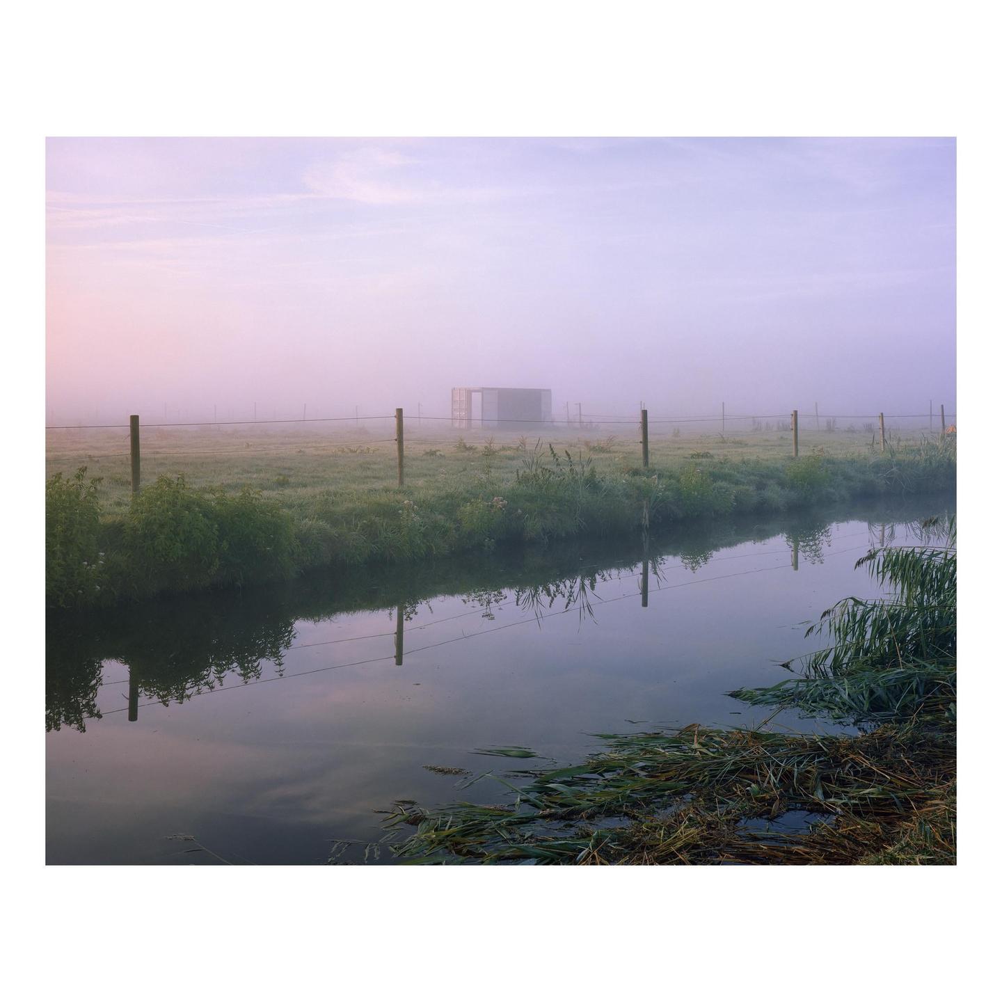 A misty field with a fence and a canal in the foreground, and a building in the distance.