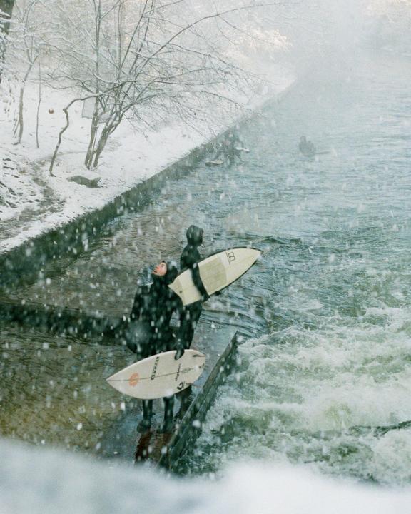 Two surfers in wetsuits stand on the bank of a river holding their surfboards while it snows.