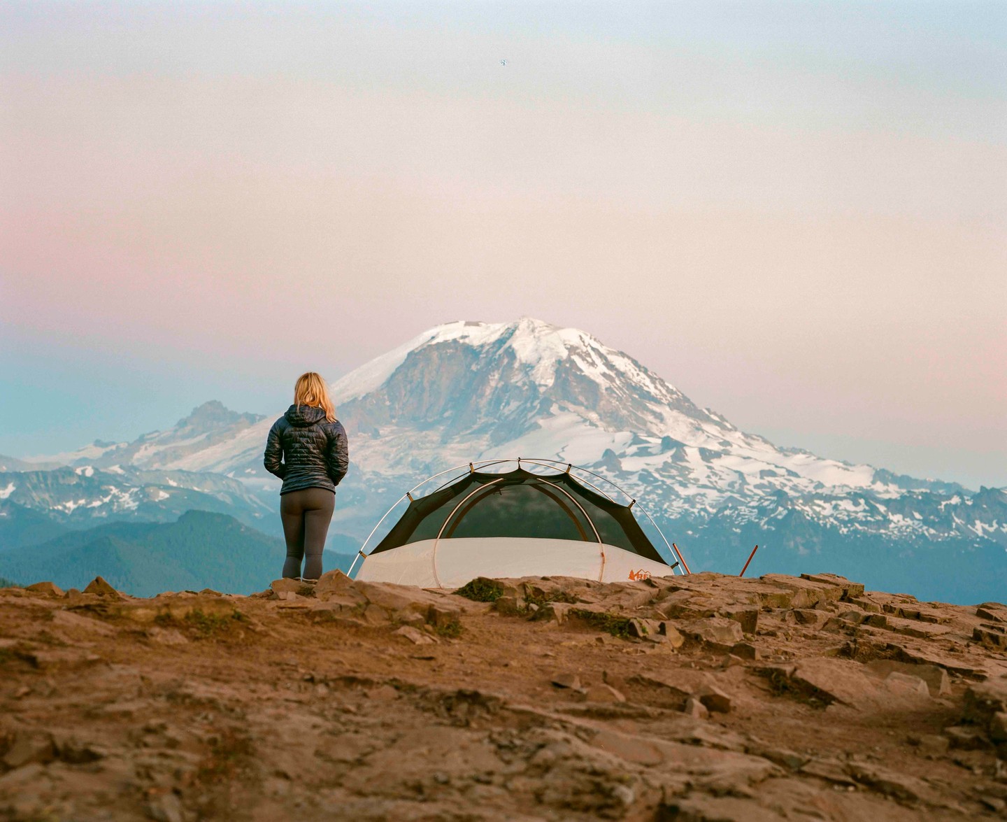 A woman stands next to a tent on a rocky outcrop, looking out at a snow-capped mountain range under a pastel sky.