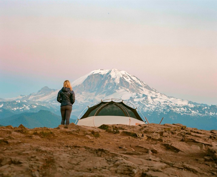 A woman stands next to a tent on a rocky outcrop, looking out at a snow-capped mountain range under a pastel sky.