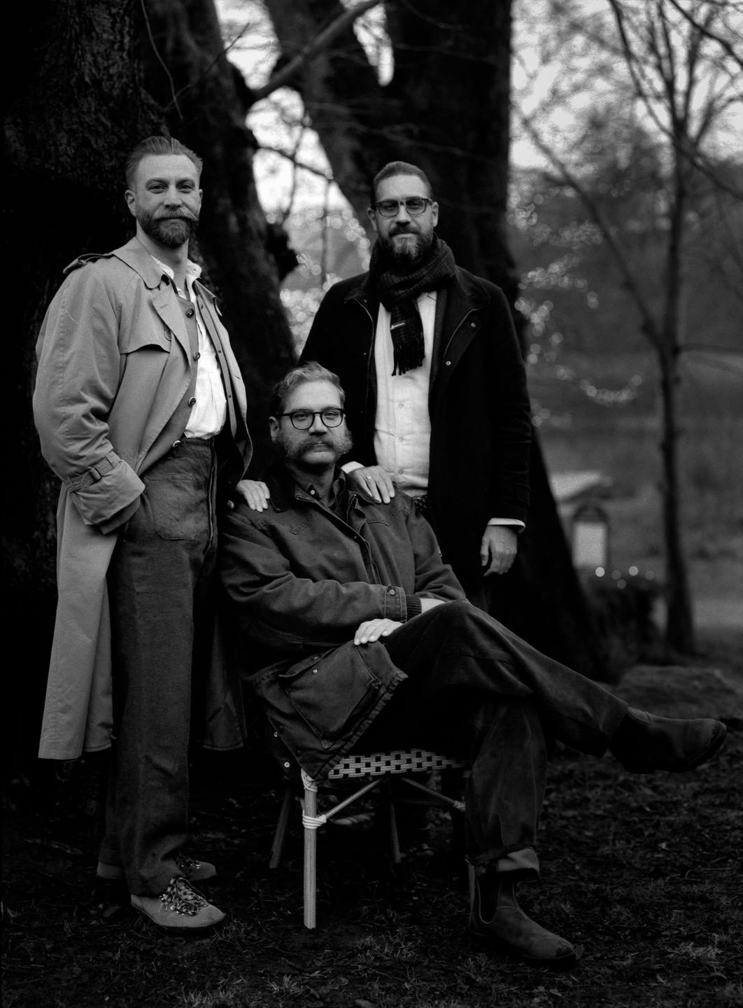 Three men with beards and mustaches pose outdoors, with one man seated and the other two standing behind him.