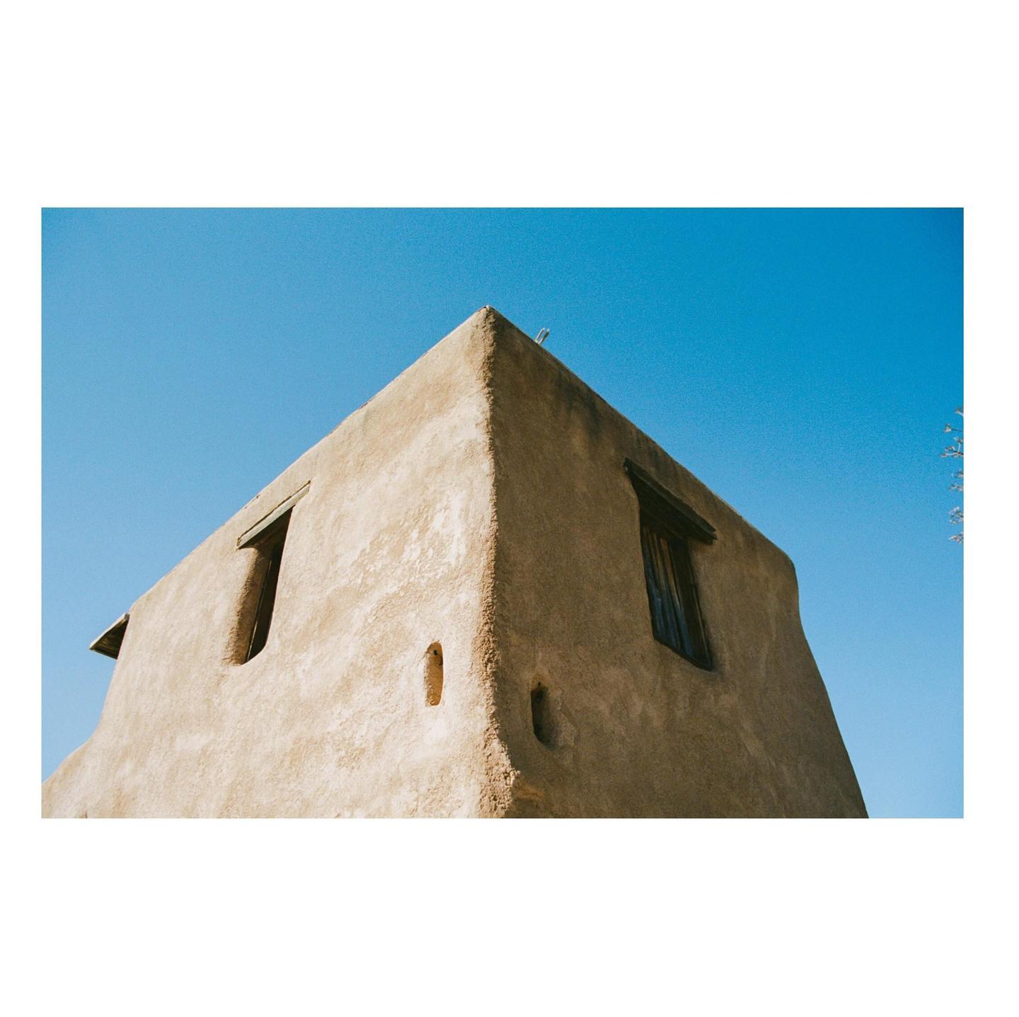 A corner of a textured adobe building with small windows is shown against a clear blue sky.