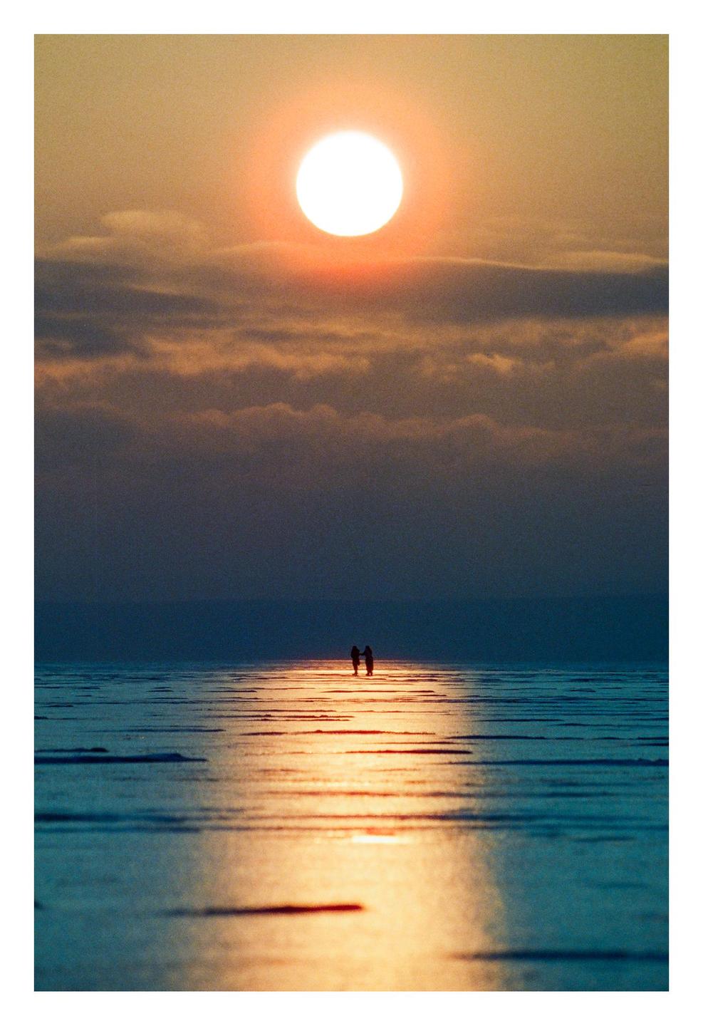 A couple stands on a frozen lake with the setting sun reflecting on the ice.