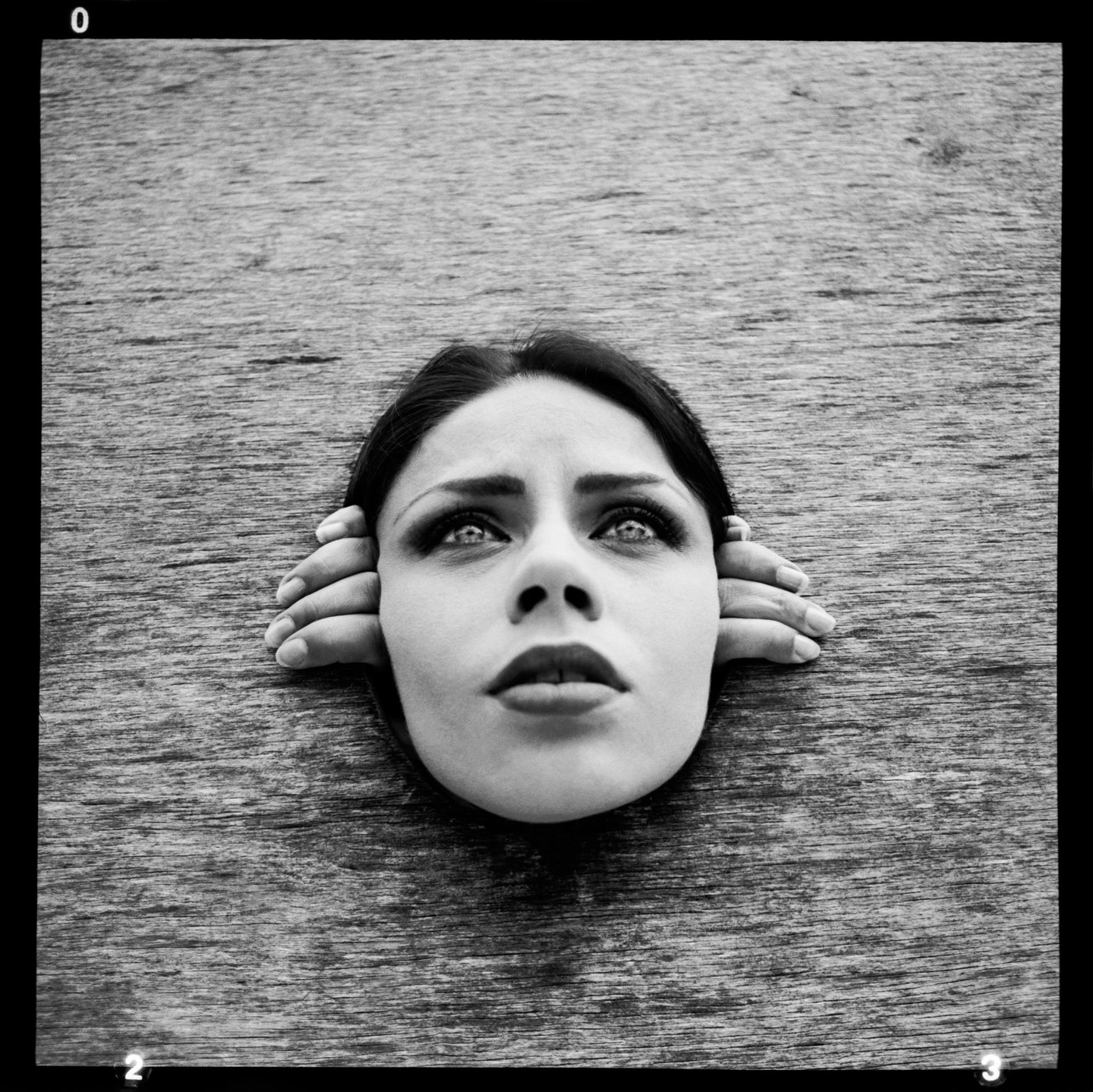 A woman's head and hands emerge from a wooden surface, looking upwards.