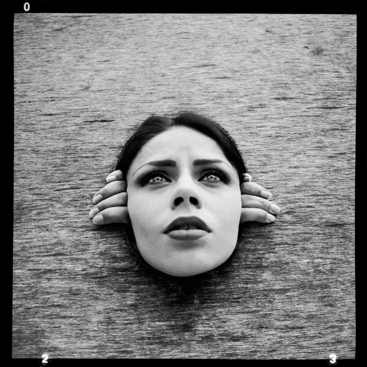 A woman's head and hands emerge from a wooden surface, looking upwards.
