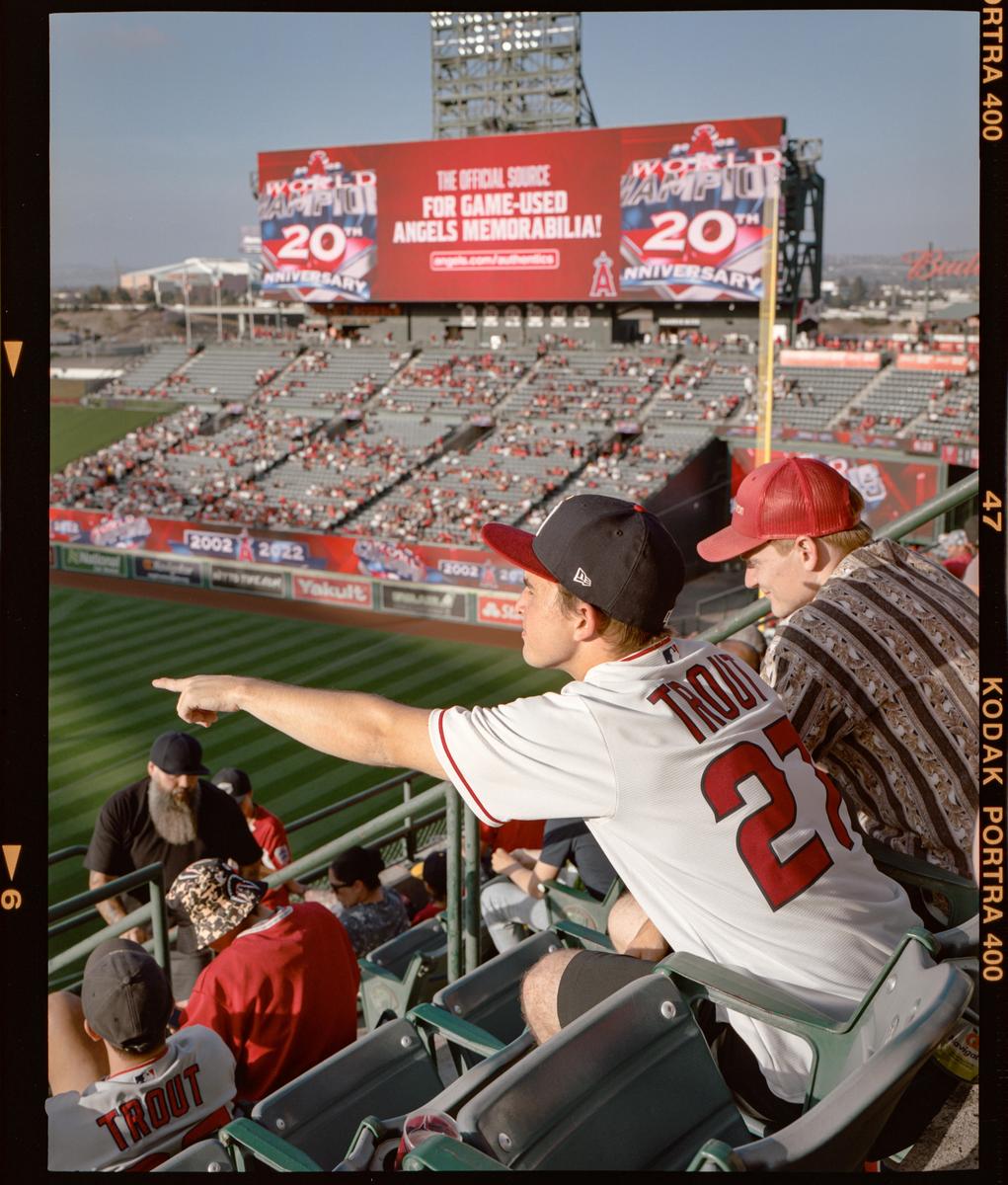 A man in a baseball jersey points towards the field from the stands of a baseball stadium.