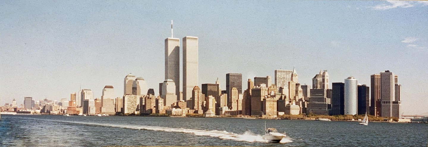 A boat creates a wake on the water in front of the New York City skyline with the Twin Towers visible.