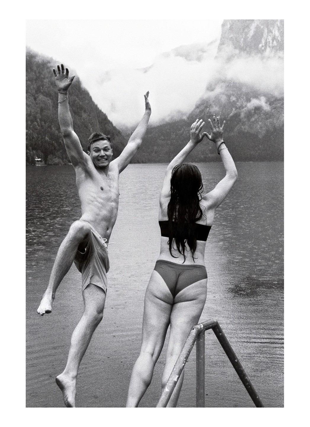 A man jumps into a lake next to a woman standing on a metal ladder, with mountains and clouds in the background.