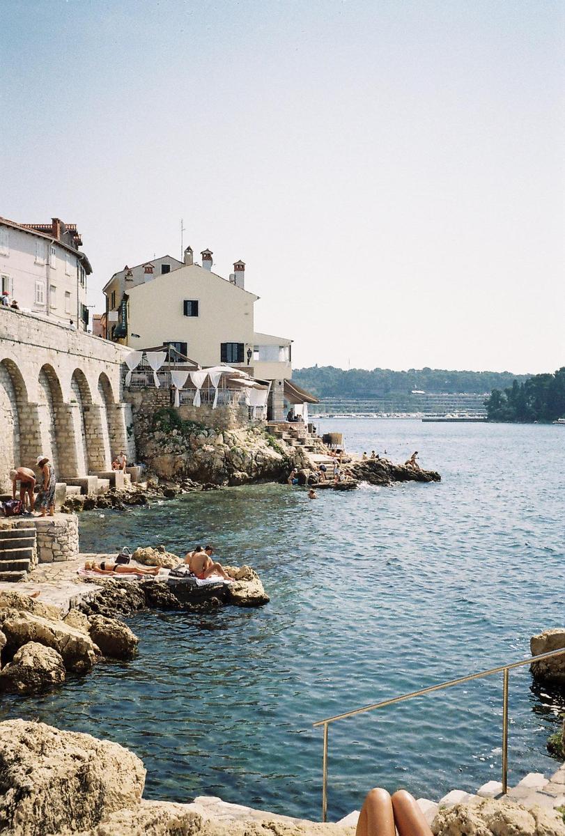 People are sunbathing and swimming in the sea next to buildings built on a rocky shore.