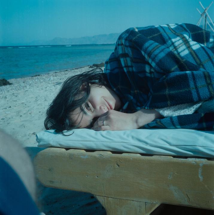A woman wrapped in a plaid blanket lies on a wooden lounge chair on a sandy beach with the ocean and mountains in the background.