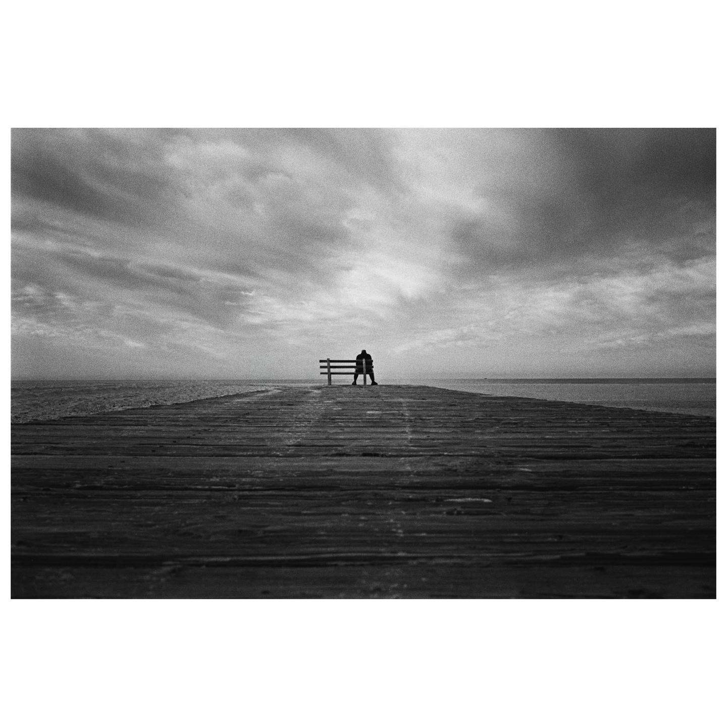 A person sits on a bench at the end of a pier under a cloudy sky.