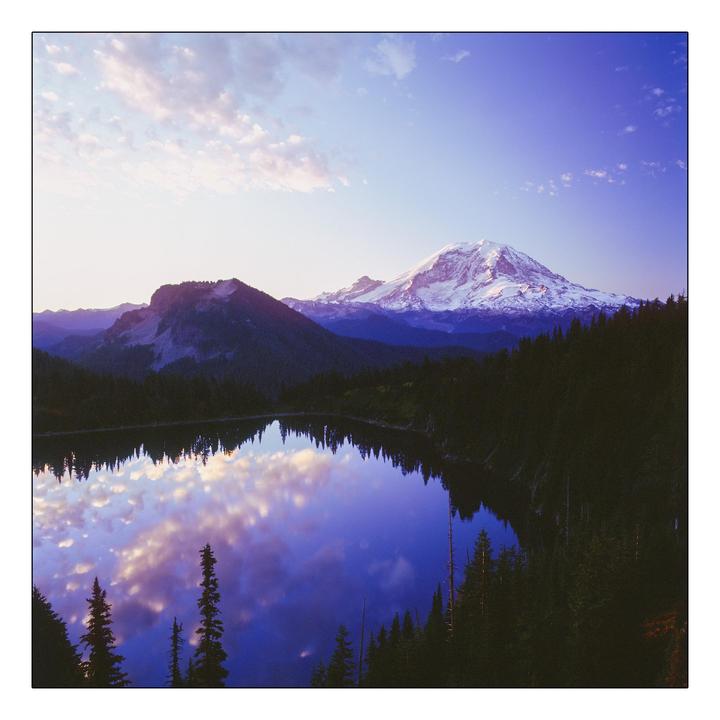 A snow-capped mountain and forest are reflected in a lake under a cloudy sky.