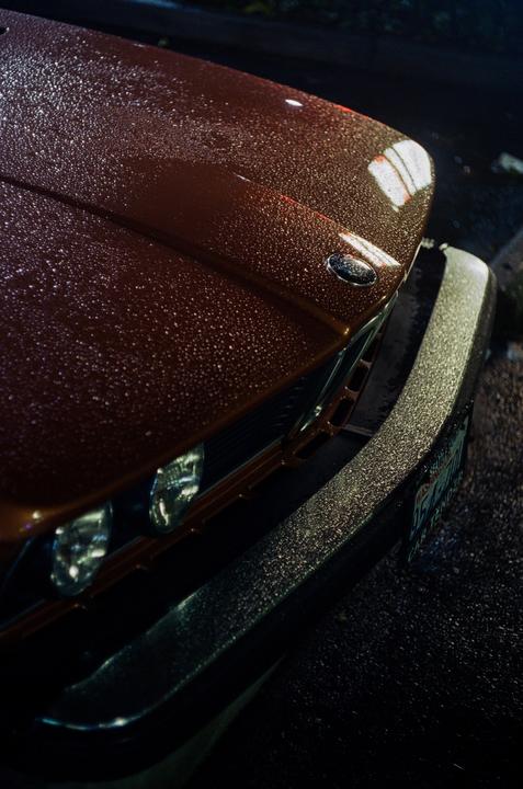 Raindrops cover the hood and bumper of a brown car at night.