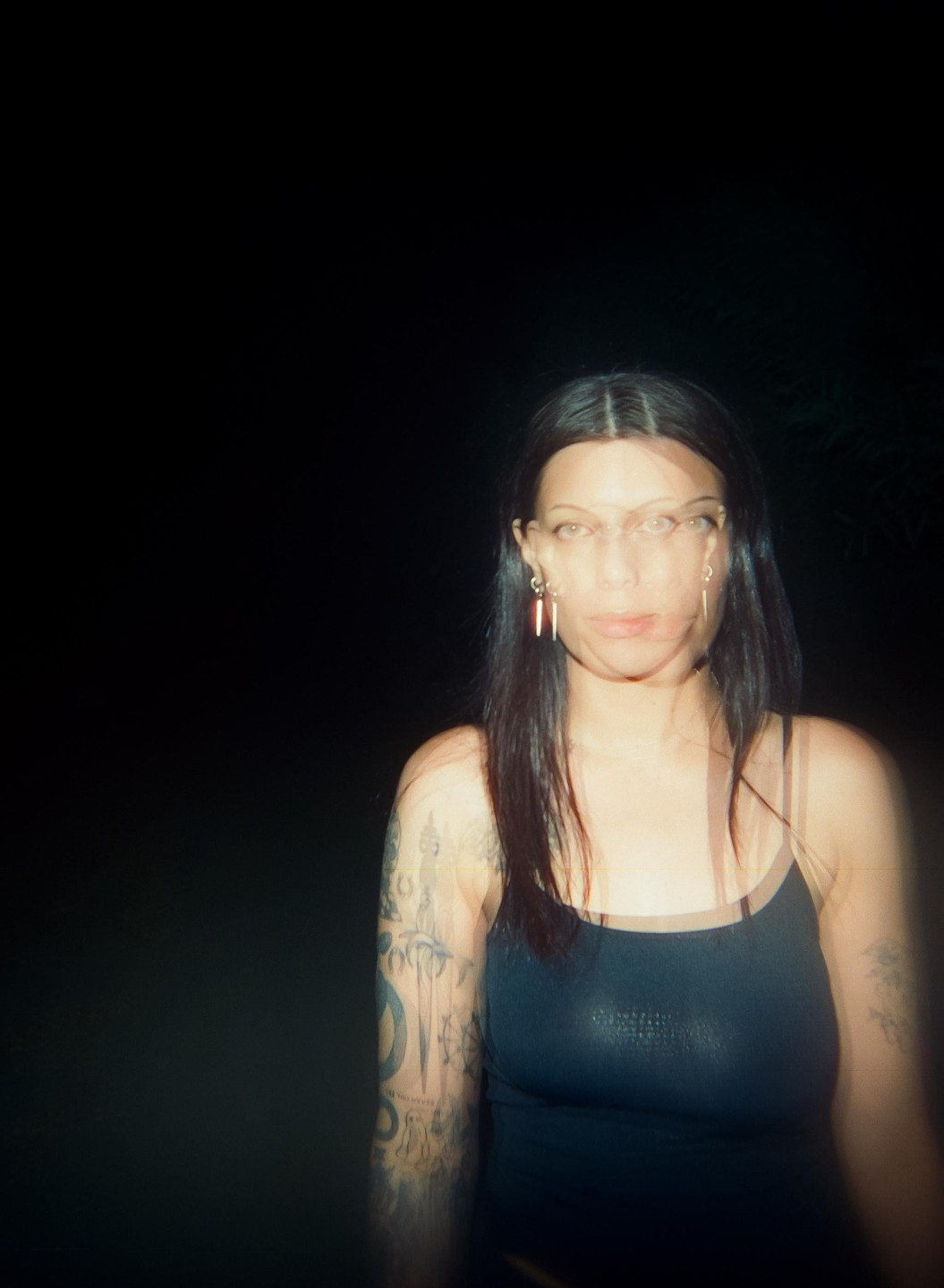 A woman with long dark hair and tattooed arms looks forward in a blurry night portrait.