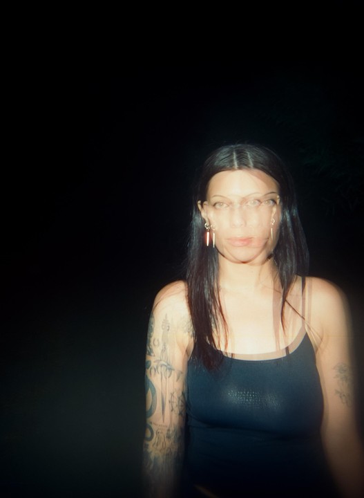 A woman with long dark hair and tattooed arms looks forward in a blurry night portrait.