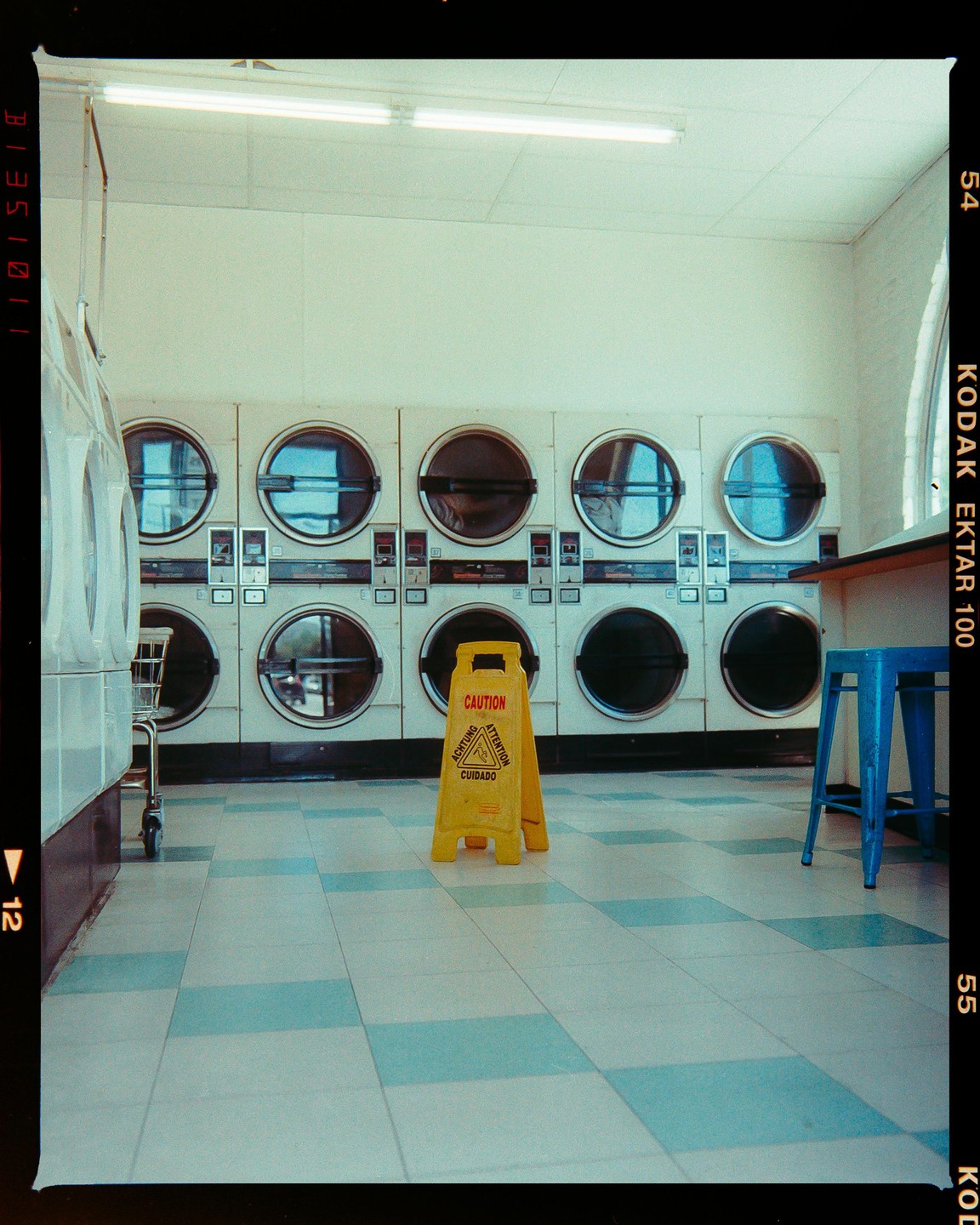 A yellow caution sign stands on a tiled floor in front of a row of washing machines.