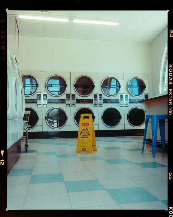 A yellow caution sign stands on a tiled floor in front of a row of washing machines.