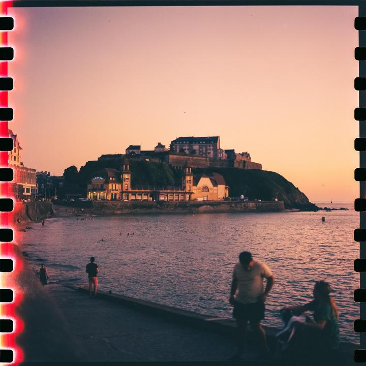 A coastal town with buildings on a hill overlooks a body of water where people are swimming and walking along the shore at sunset.