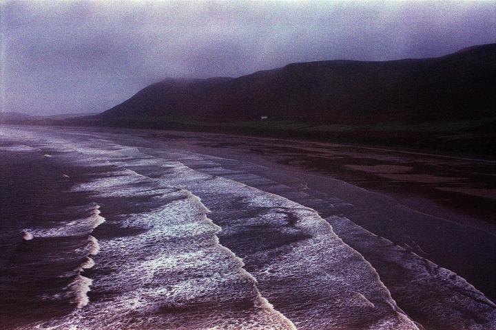 Waves crash on a beach with a mountain in the background under a hazy sky.